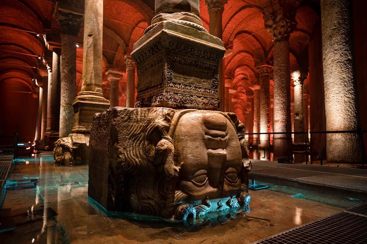 Lit marble columns and a carved Medusa head reflected in still water in the Basilica Cistern